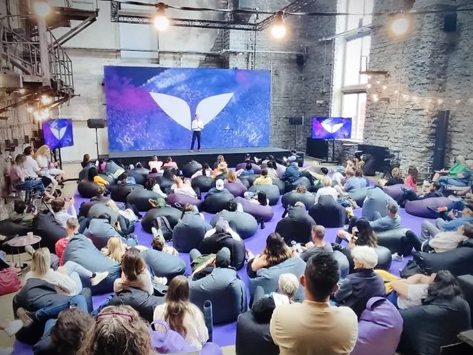 A large audience seated on bean bags in a spacious industrial-style conference room, listening to a speaker on stage with a large screen displaying a logo. The room features exposed brick walls and high windows.