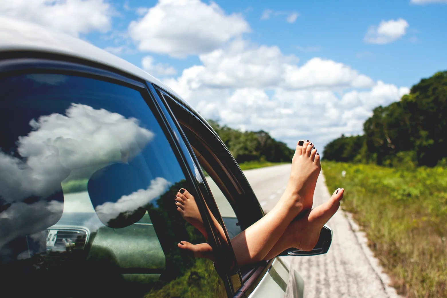Bare feet resting out of a car window on an open road lined with greenery