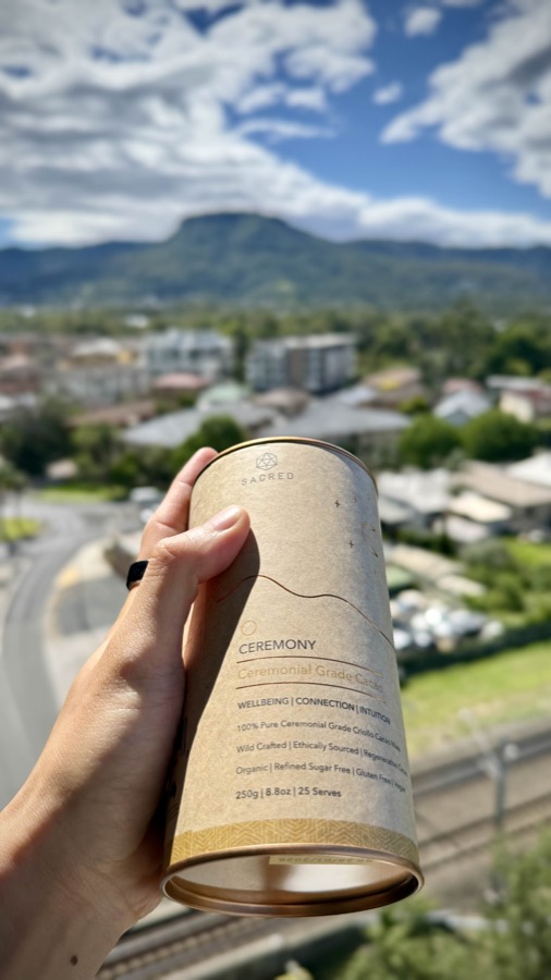 Sacred Taste ceremonial-grade cacao tin held up with a mountain view behind